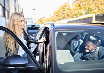 A woman entering a dark gray car through the open passenger car door while the male driver smiles and watches her.