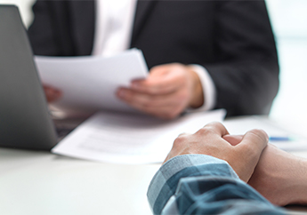 A person sitting with their hands folded together on a table, while another person reviews paperwork while sitting in front of a laptop computer.