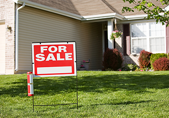 A red "For sale" sign in the front yard of a beige home.