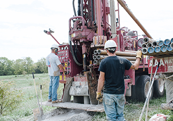 A well driller wearing a hardhat standing on the back platform of a red well drilling rig
