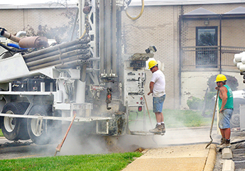 A well driller wearing a hardhat standing on the back platform of a well drilling rig while actively drilling a well
