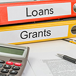 Two 3-ring binders on a table, an orange binder marked "loans" and a yellow marked "Grants", with a calculator, pen and paper copy of terms in front of the binders 
