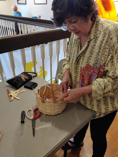 Woman standing at a table with scissors and a screwdriver on it near a railing, weaving a basket.