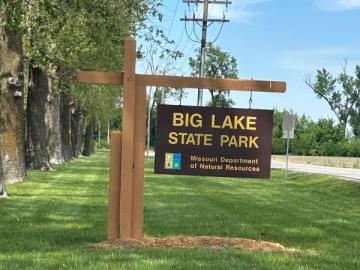 Brown cantilever sign with Big Lake State Park, MO Dept of Natural Resources. Trees line the left side, and sign is near the road on a grassy area.